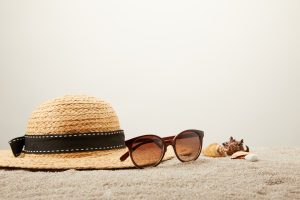 vue rapprochée d'un chapeau de paille, de lunettes de soleil et de coquillages sur le sable sur fond gris